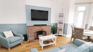 Un salon avec une cheminée en briques, un téléviseur à écran plat fixé au-dessus, un fauteuil en osier, un canapé bleu et une table basse blanche.