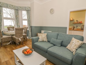 A cozy living room with a green sofa, wicker chairs, a dining table with glasses, and a bowl of oranges on a white table.