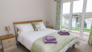 A bedroom with a bed, two bedside tables, a window with curtains, and a view of the outside. - Mor Awelon (Goodwick)