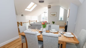 A dining area with a wooden table set for four, grey upholstered chairs, and a view of a living room with a sofa and a painting on the wall. - Mor Awelon (Goodwick)
