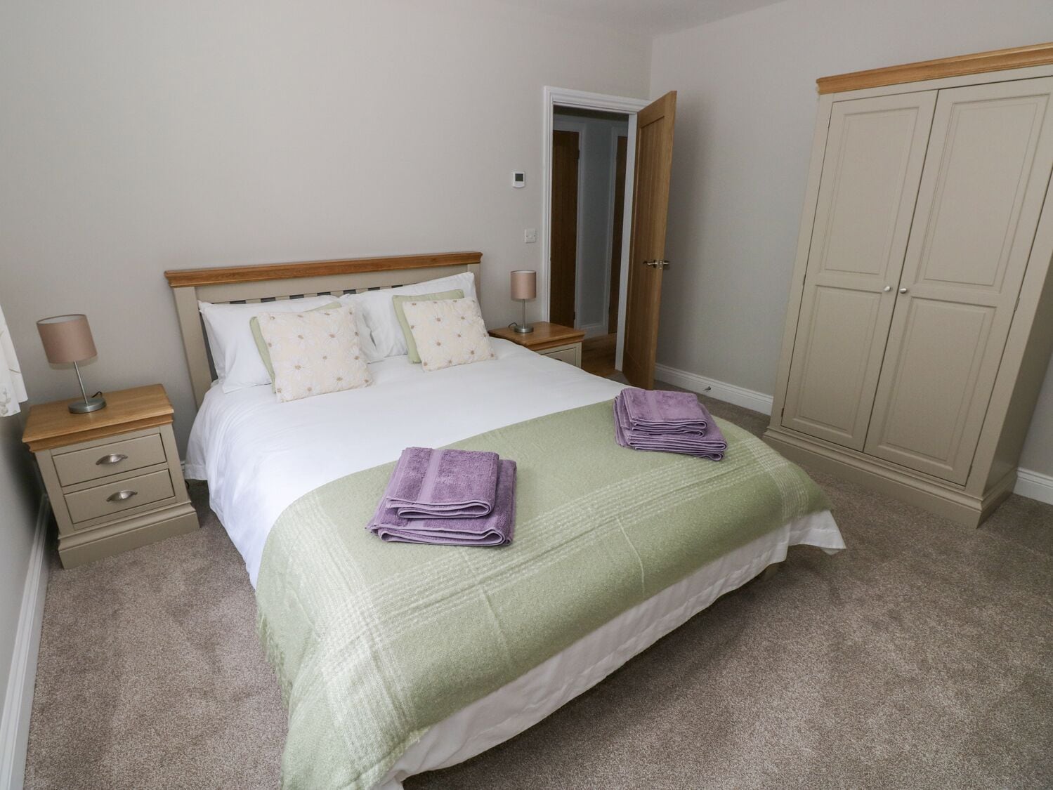 A neatly made bed with a green bedspread and two folded purple towels. There is a wooden nightstand with a lamp and a drawer. A wardrobe is visible in the background.