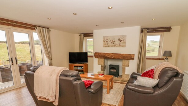 A spacious living room with two leather sofas, a wooden coffee table, a flat-screen TV, and a stone fireplace. - Meadow Cottage (Keighley)