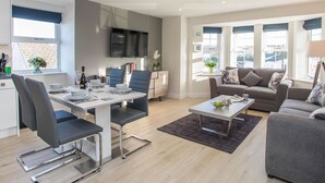 Un salon moderne comprenant un coin repas, des canapés gris, un téléviseur à écran plat et de grandes fenêtres.