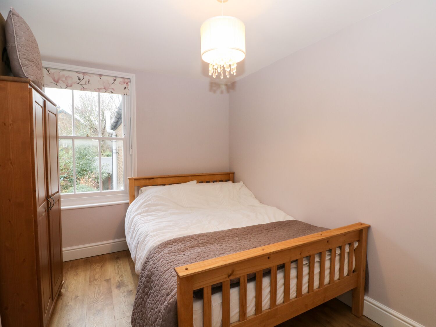 A bedroom with a wooden bed, a window with floral curtains, a wooden wardrobe, and a hanging light fixture.
