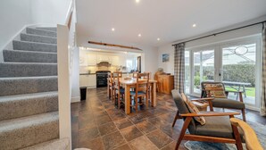 A spacious living area with a dining table and chairs, a staircase, and a kitchen in the background. - Curragho (Holyhead)