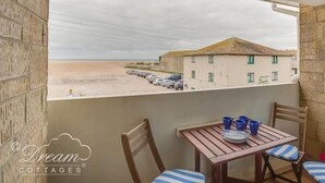 Un balcon avec une table et des chaises, donnant sur une plage et un immeuble où sont garées des voitures.