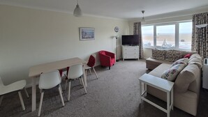 Un salon comprenant un canapé, un téléviseur, une table à manger avec des chaises et une fenêtre donnant sur la plage.