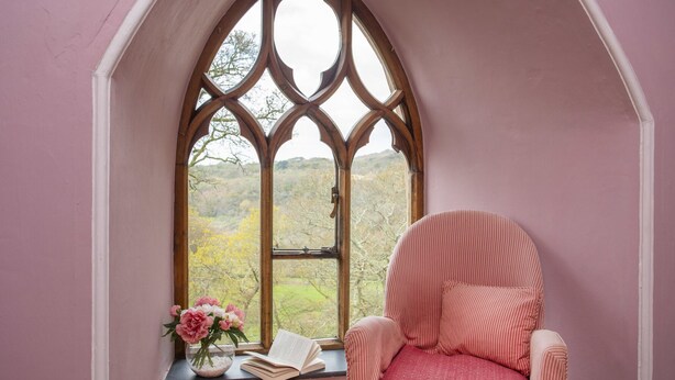 A room with a gothic window, a chair, a vase with flowers, and an open book on a windowsill. - Brynkir Tower (Garndolbenmaen)