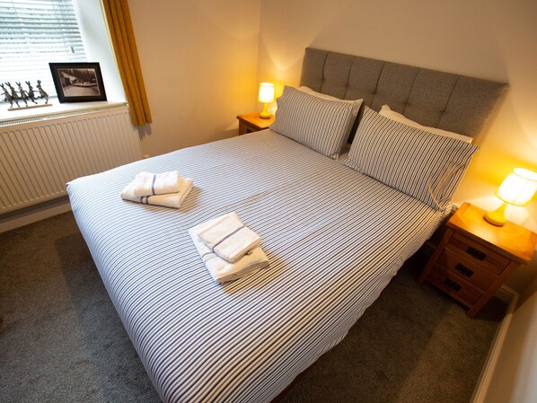 A neatly made bed with striped bedding and two folded towels, bedside tables with lamps, a radiator, and a window with blinds and a picture frame.