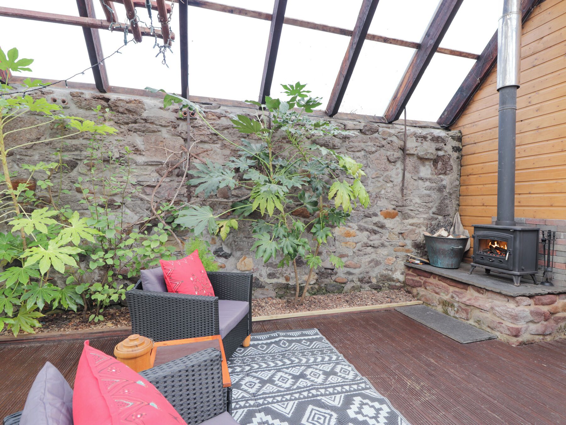 Un espace extérieur accueillant, aménagé avec des meubles en osier et un tapis à motifs, un mur en pierre et un poêle à bois.