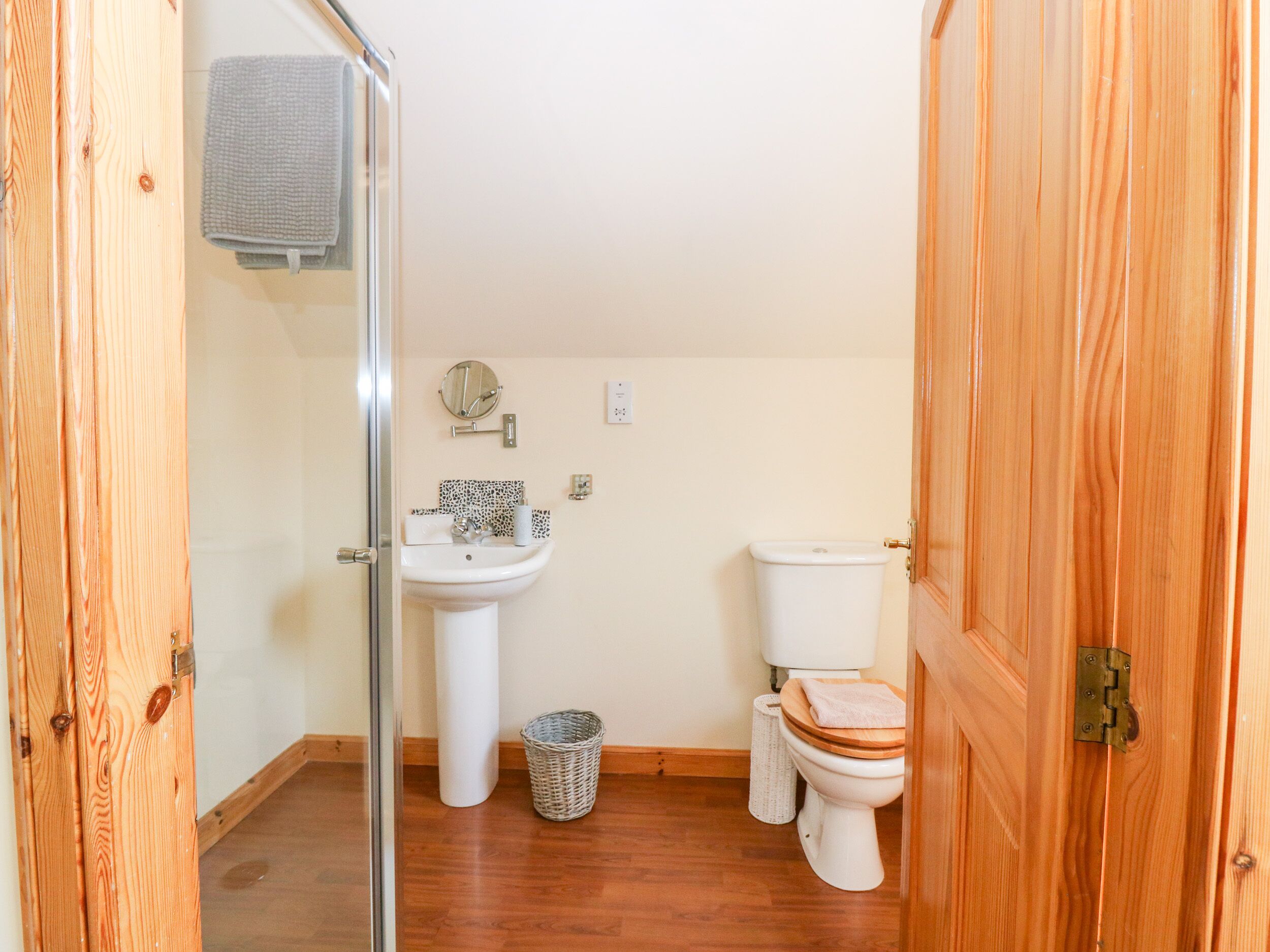 A small, clean bathroom with a wooden floor, white fixtures, and a glass shower enclosure.