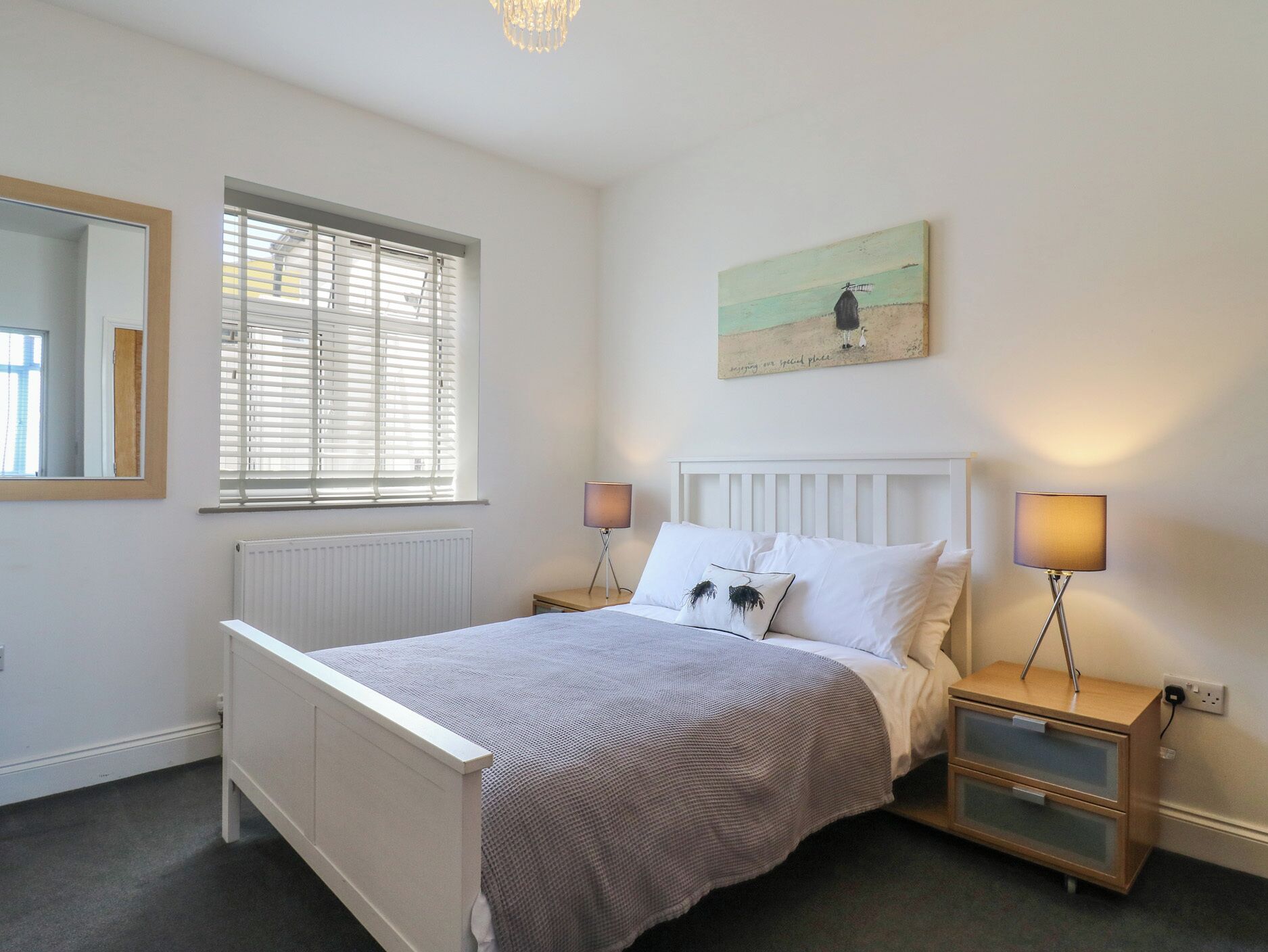 Une chambre à coucher avec un lit, des tables de chevet, un miroir, une fenêtre avec des stores et un tableau au mur.