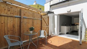 Um deck de madeira com duas cadeiras e uma mesa, uma porta de correr de vidro que dá para uma cozinha e um cercado de madeira.