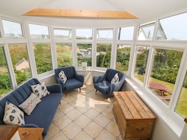 Uma sala de sol com vista para um jardim e casas. Possui um sofá azul, um baú de madeira e azulejos hexagonais.
