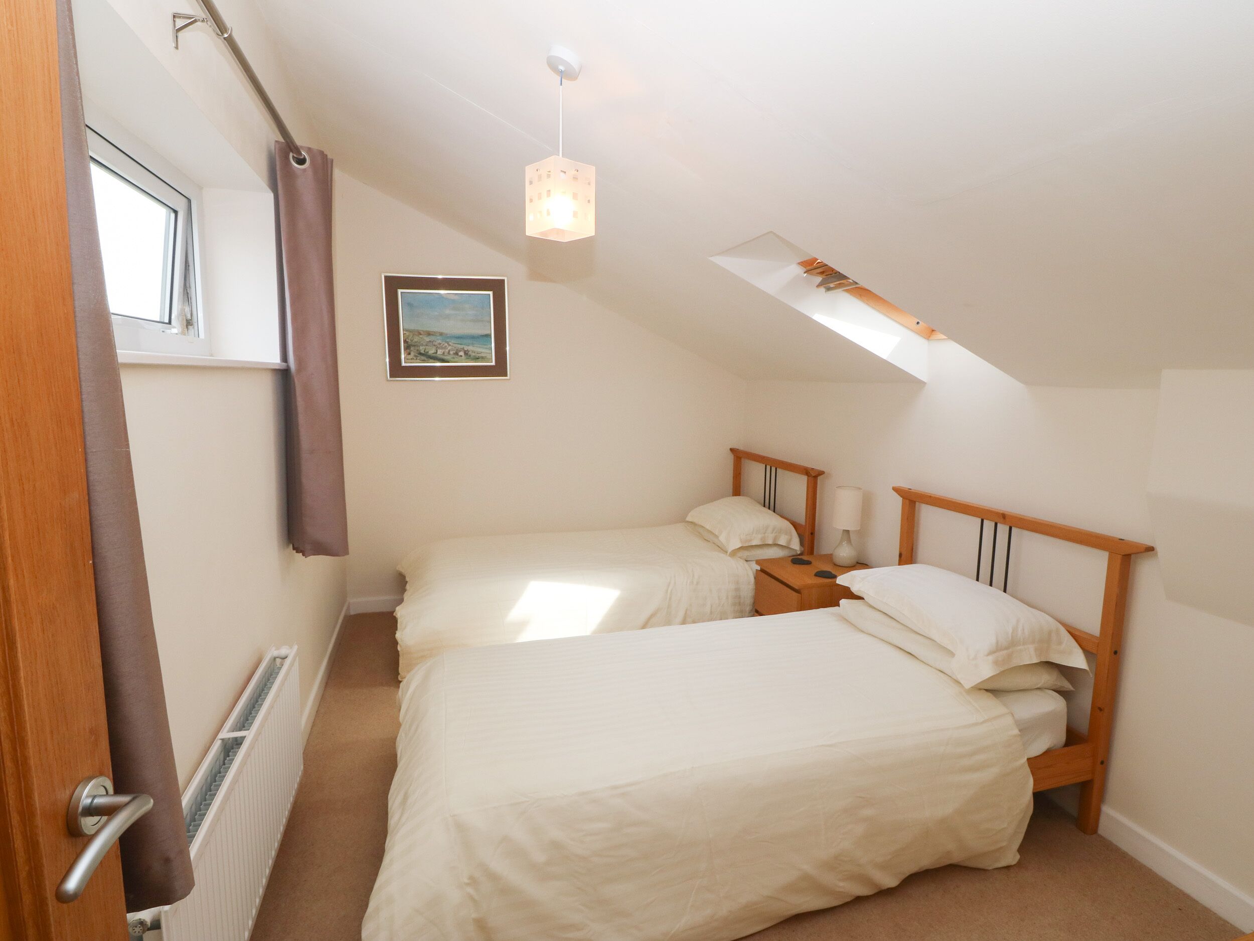 A small, well-lit bedroom with two single beds, a nightstand, a lamp, and a framed picture on the wall.