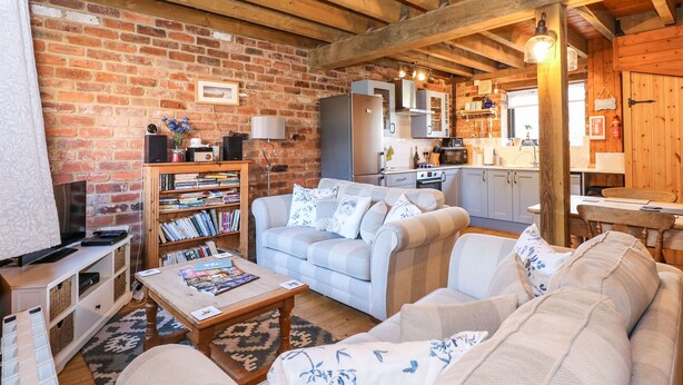 A cozy living room with a fireplace, a bookshelf, a sofa, and a dining area with wooden furniture. - The Dairy Barn (Fakenham)