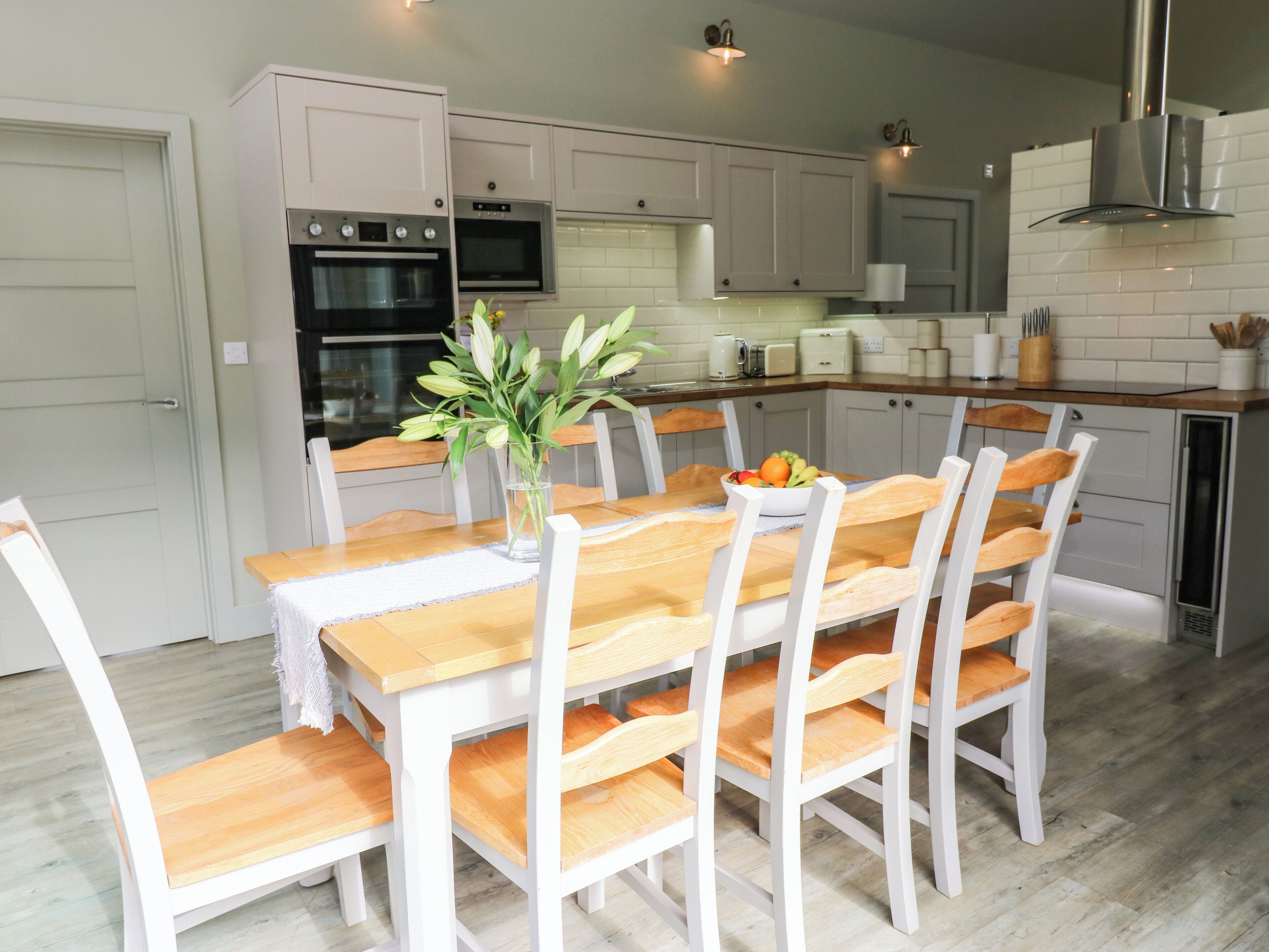 A modern kitchen with white cabinetry, a wooden dining table, and chairs. There is a vase with flowers on the table.