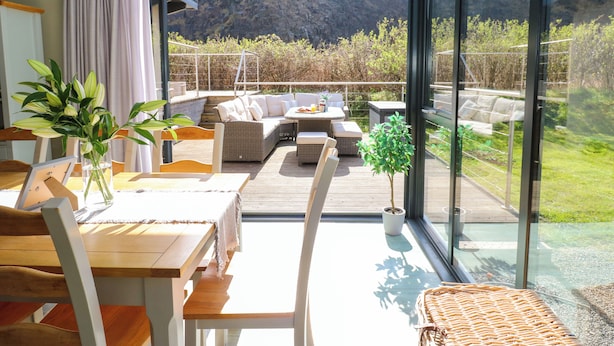 A modern outdoor living area with a wooden deck, wicker furniture, and a view of a mountainous landscape. - Restharrow Lodge (Bamburgh)