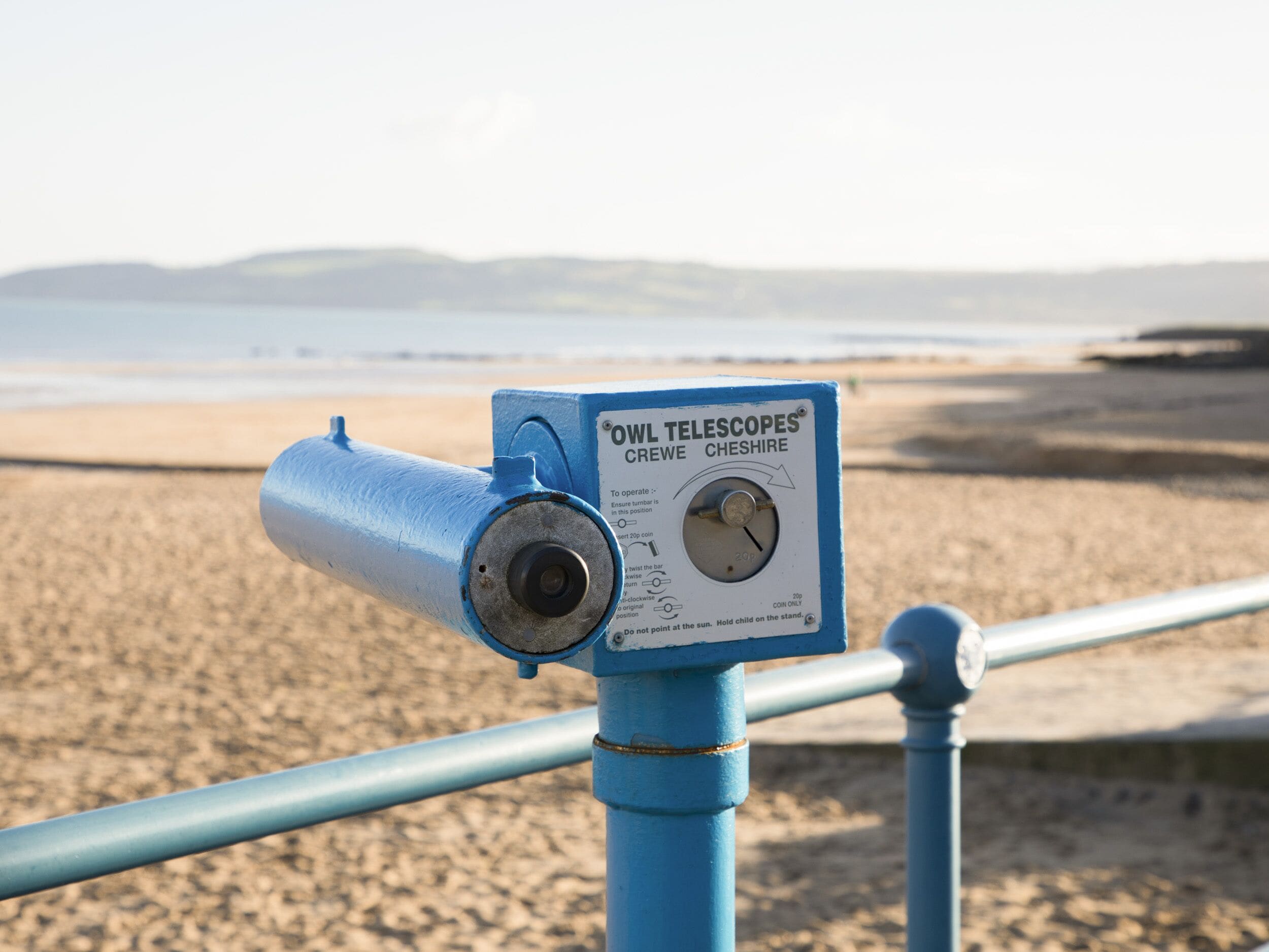 A telescope labeled "OWL TELESCOPES CREWE CHESHIRE" mounted on a blue metal pole at a beach.