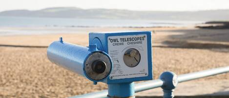 A telescope labeled "OWL TELESCOPES CREWE CHESHIRE" mounted on a blue metal pole at a beach.