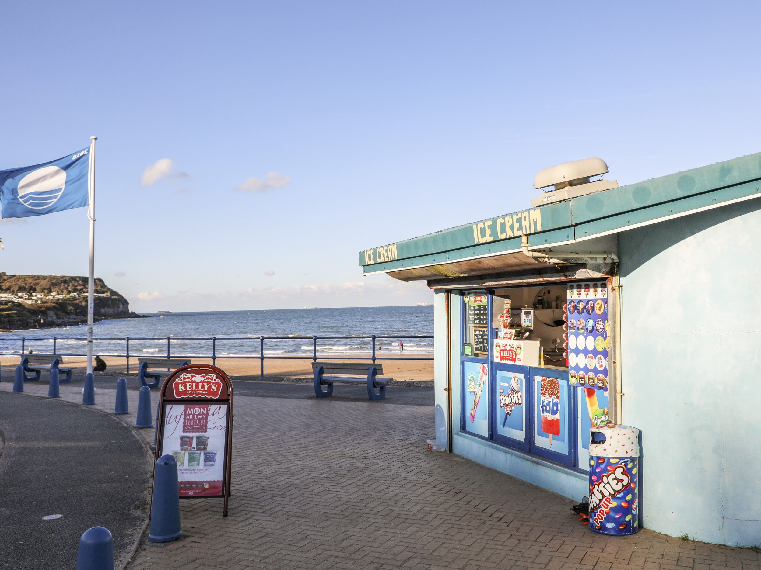 A beachside ice cream cart with a sign reading "ICE CREAM" and a nearby "KELLY'S" signboard.
