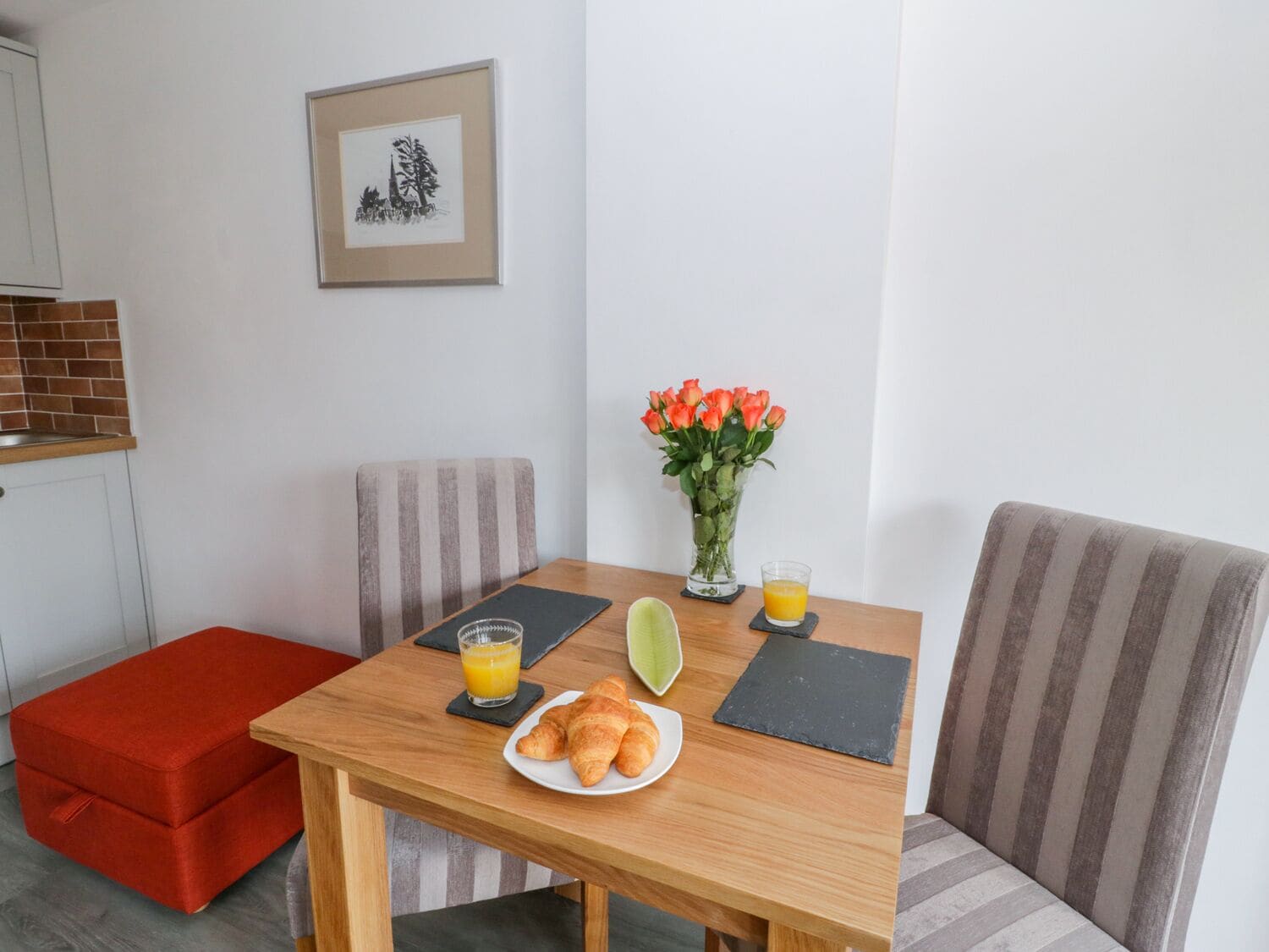 Un comedor con mesa de madera, cojines a rayas, un reposapiés rojo, un cuadro en la pared, un jarrón con flores y un plato de croissants.