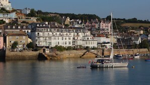 A coastal town with a marina, featuring a large white building with multiple balconies, a small boat docked nearby, and a clear blue sky. - 3 Melbury (Salcombe)