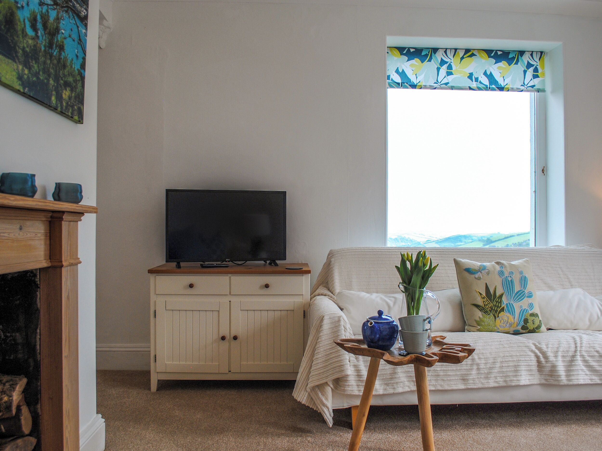 Ein Wohnzimmer mit Fernseher auf einem Holzständer, ein weißes Sofa mit floralen Kissen, ein kleiner Holztisch mit einer Vase und einem Teekessel sowie ein Fenster mit einem blumigen Vorhang.