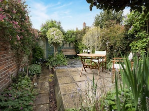 A garden patio with a wooden table and chairs, surrounded by lush greenery and flowering plants.
