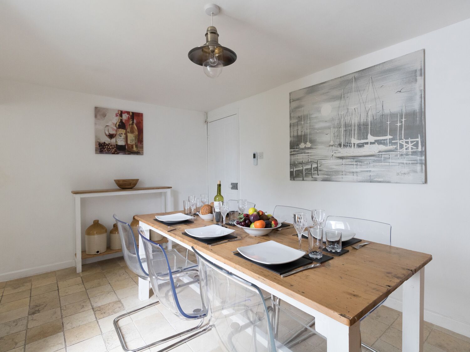 A dining area with a wooden table set for four, clear chairs, a bowl of fruit, wine glasses, and a bottle of wine.
