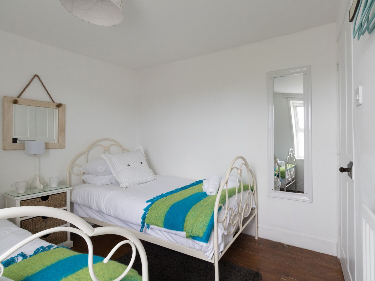 A bedroom with a white metal bed frame, white bedding, a green and blue striped blanket, a bedside table with a lamp, and a mirror on the wall.