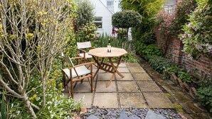 A patio with a round wooden table and two striped chairs, surrounded by lush greenery and a brick wall. - Fenders (Weymouth)