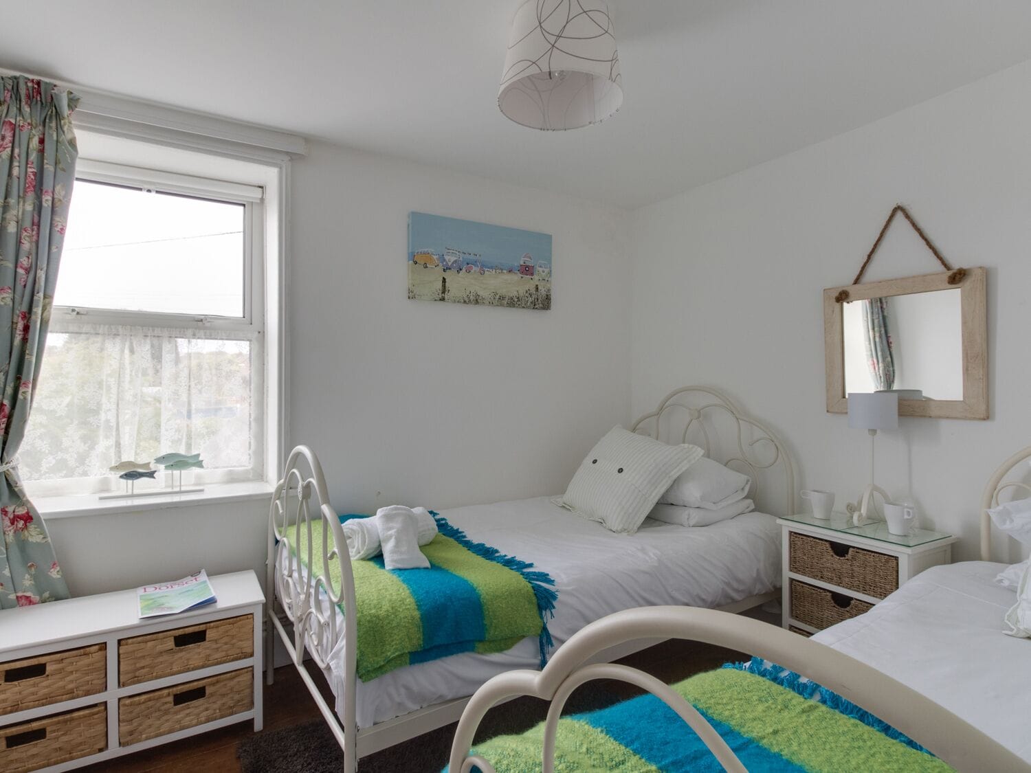 A bedroom with two single beds, a window with curtains, a bedside table with a lamp, a wall-mounted mirror, and a painting on the wall.