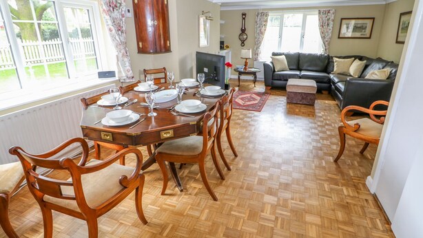 Un coin repas avec une table en bois dressée pour six personnes, entourée de chaises en bois. Un canapé noir et une petite table avec une lampe se trouvent à l’arrière-plan.