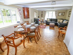 Un coin repas avec une table en bois dressée pour six personnes, entourée de chaises en bois. On aperçoit un canapé et une petite table avec une lampe à l’arrière-plan.