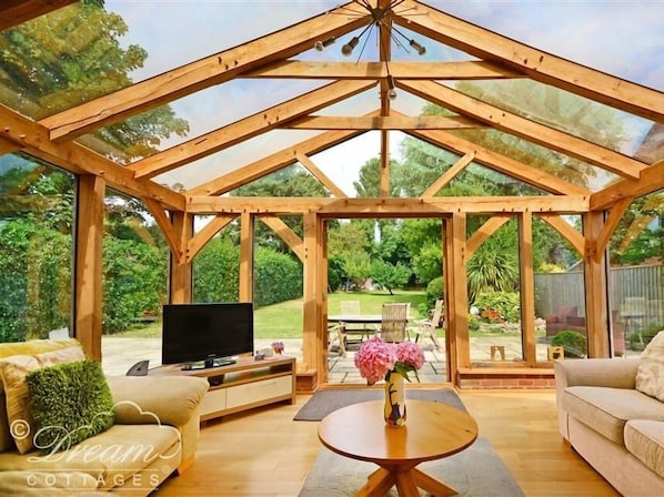 A spacious wooden conservatory with a TV, sofa, and table, overlooking a lush garden.