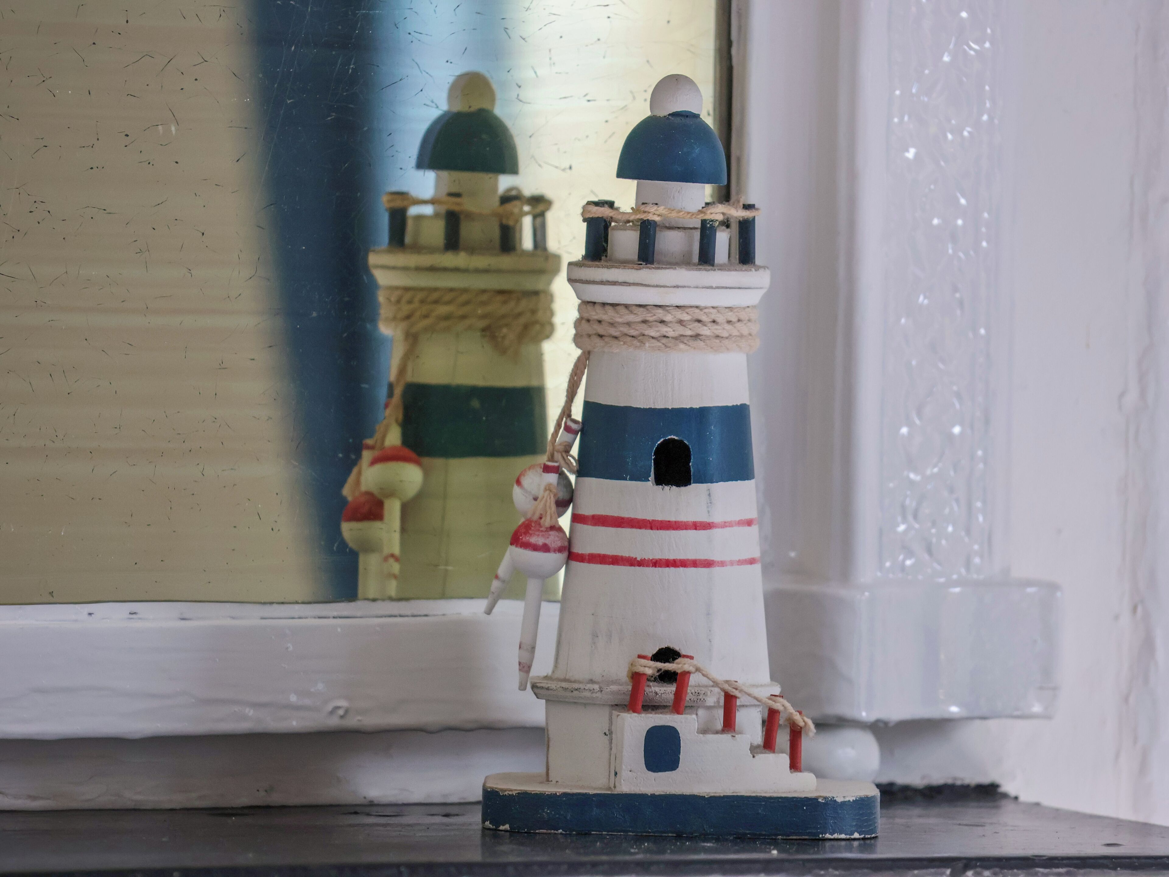 A miniature lighthouse model with a rope wrapped around its base, a small figure of a person, and a reflection of a similar lighthouse in a mirror.