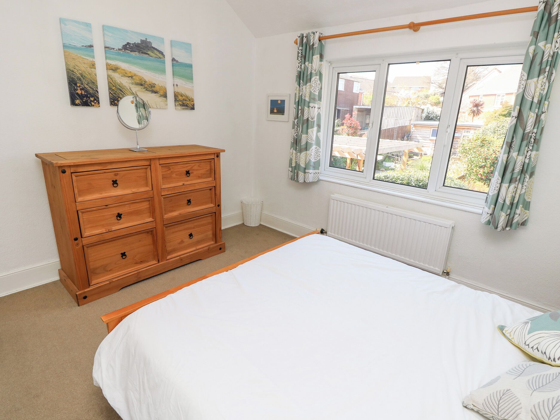 A bedroom with a wooden dresser, a bed with white bedding, a window with curtains, and beach-themed wall art.