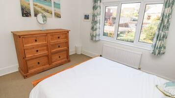 A bedroom with a wooden dresser, a bed with white bedding, a window with curtains, and beach-themed wall art.