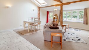 A spacious living area with a high ceiling, exposed wooden beams, a dining table with chairs, a sofa, a small wooden side table, and a large window with curtains. - The Hen House (Diss)