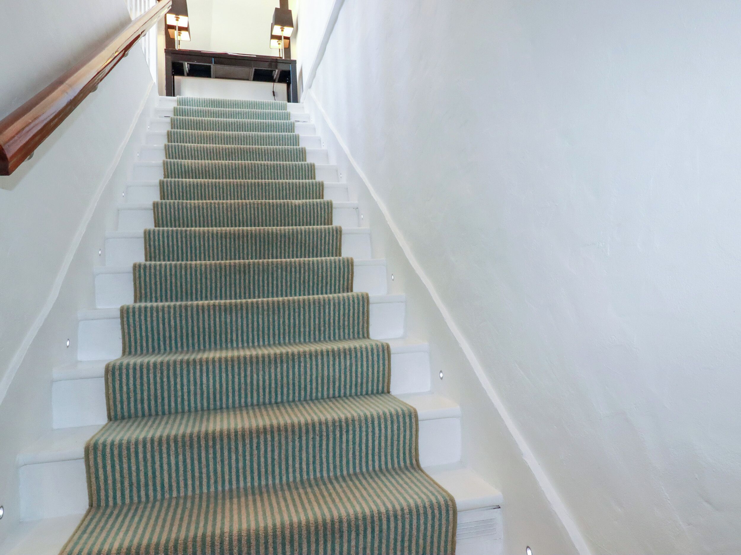 Un escalier dont les marches sont recouvertes de moquette, menant à un palier où se trouve une lampe.