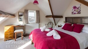 A cozy bedroom with a sloped ceiling, a wooden dresser, a chair, a bed with red bedding, and a window with curtains. - Buccaneer Cottage (Weymouth)