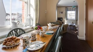 A dining area with a long table set for breakfast, a view of the city through large windows, and a cozy interior with a wooden door and an archway leading to another room. - Buccaneer Cottage (Weymouth)