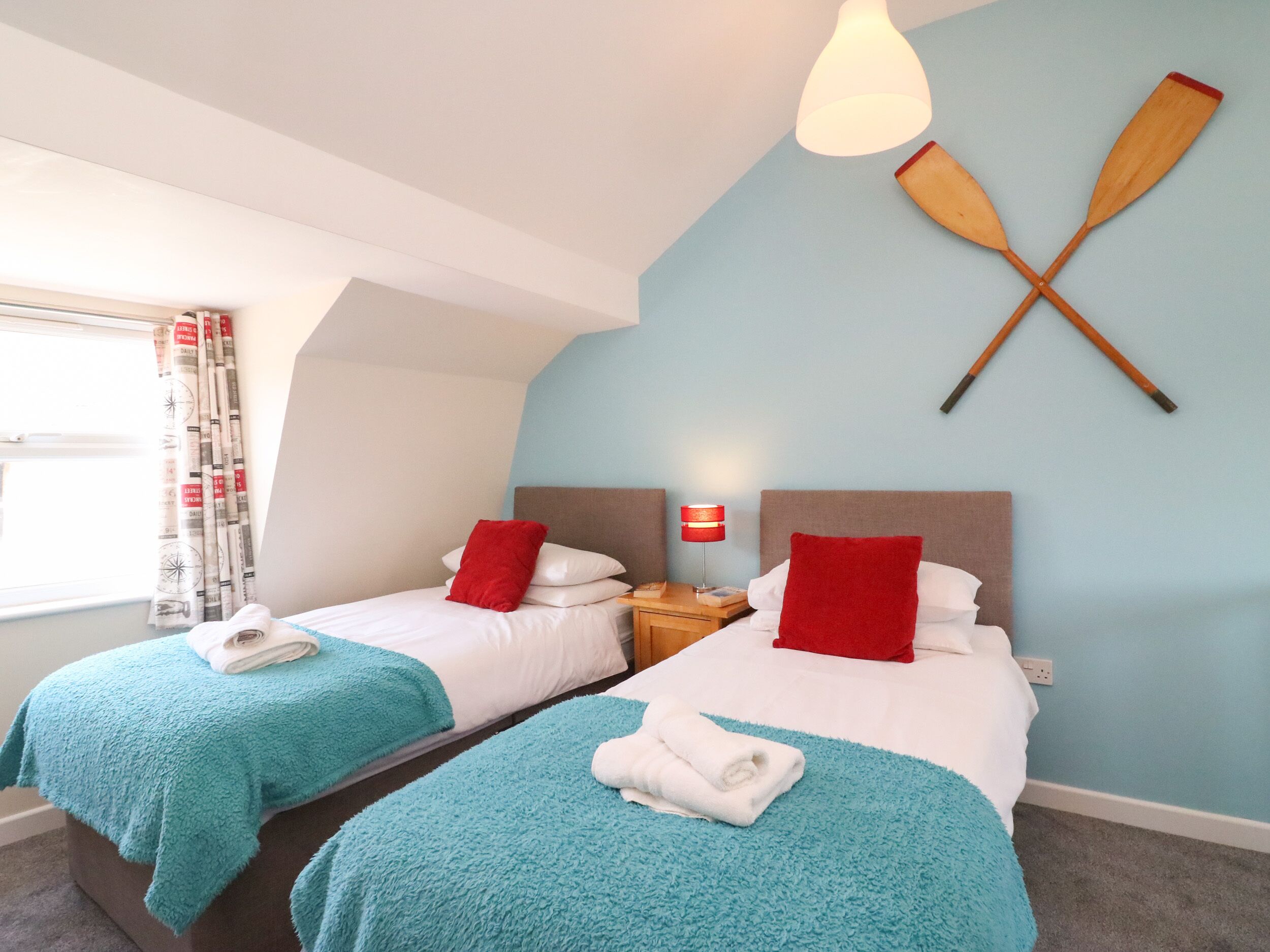 Habitación de hotel con dos camas, cada una con almohada roja y manta azul. Se observa un mesita de noche con lámpara, y dos palas de madera colgadas en la pared.