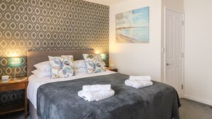 A neatly made bed with a dark grey bedspread, white pillows, and folded towels on the side. There is a patterned wallpaper on the wall behind the bed, a painting of a beach scene, and a white door to the right. - Anchor Cottage (Weymouth)