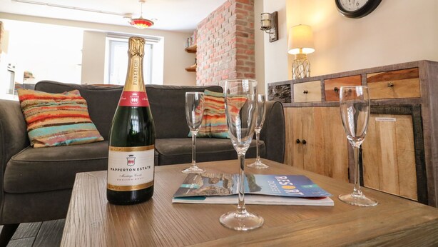A room with a wooden table, a bottle of champagne, and three wine glasses. There is a couch with colorful pillows and a brick wall in the background. - Anchor Cottage (Weymouth)
