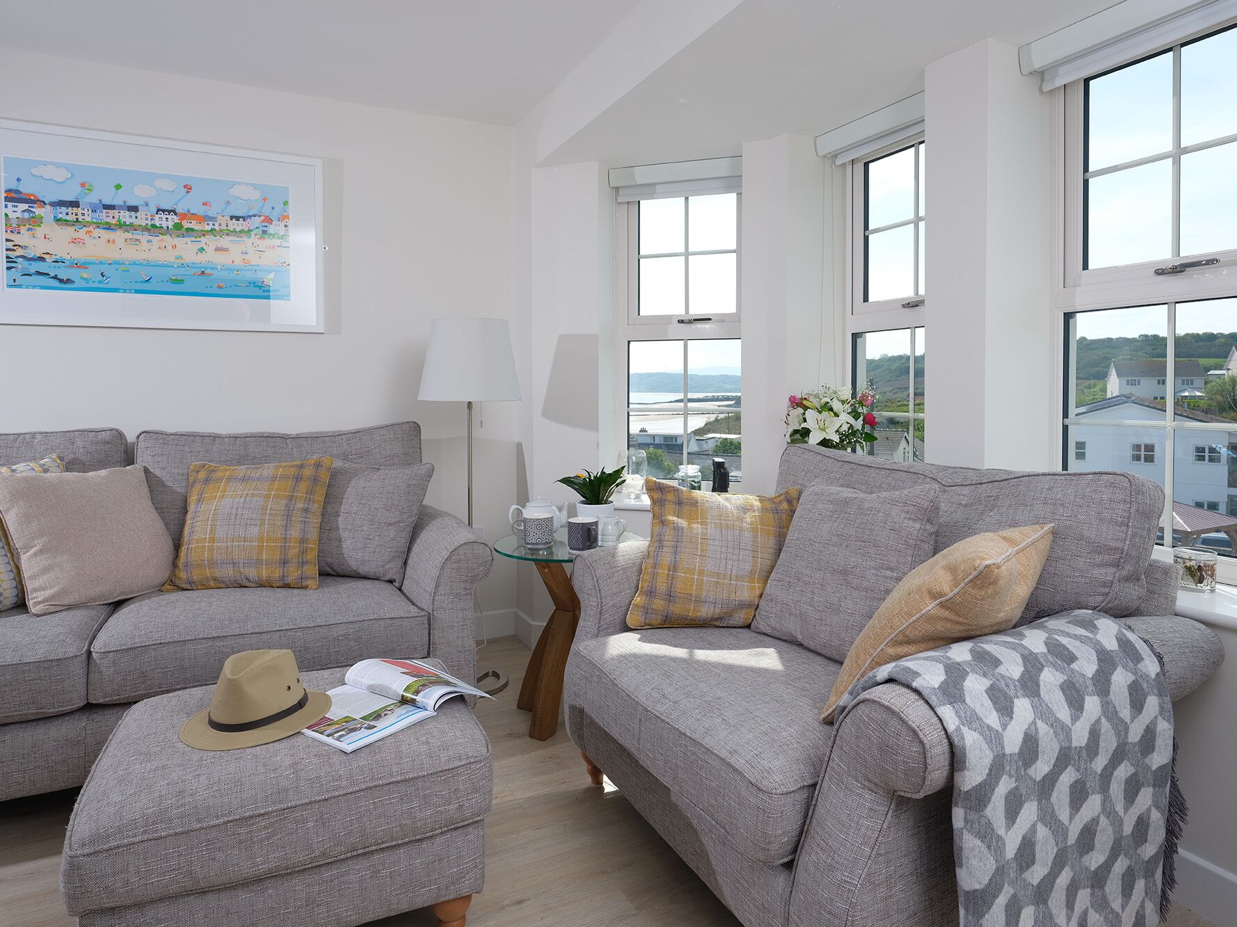 A modern living room with two grey sofas, a small coffee table, and a window with a view of the sea and buildings.