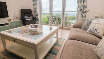 Uma sala de estar com uma mesa de centro de vidro, um sofá, uma televisão e vista para o exterior através de uma porta de correr de vidro.