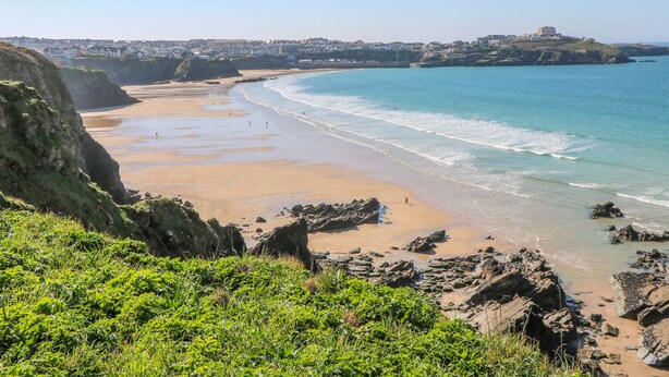 A coastal landscape with a sandy beach, rocky outcrops, and a distant building on a hill. - Ocean Seven (Newquay)