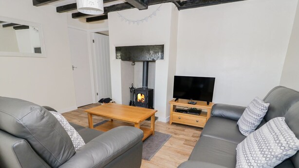 A living room with two grey leather sofas, a wooden coffee table, a TV on a wooden stand, and a wood-burning stove. - 5 Ceirnioge Cottages (Betws-Y-Coed)